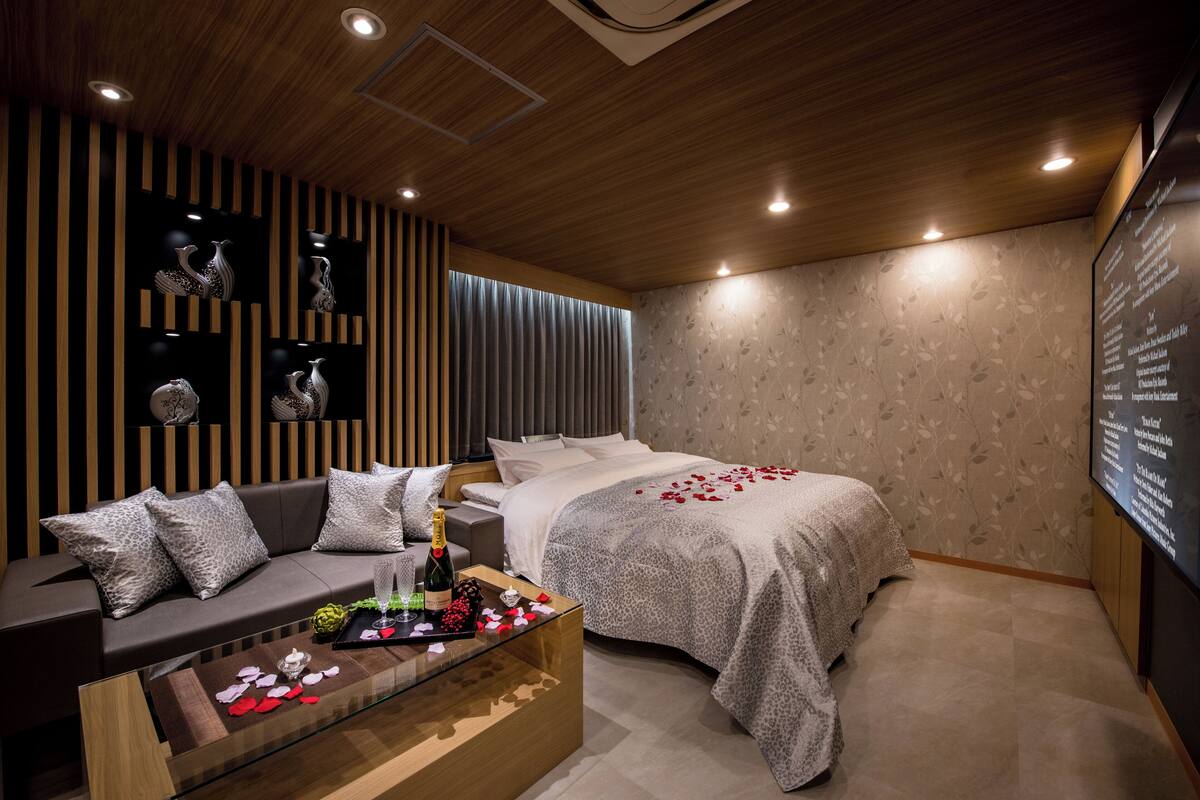 A modern bedroom with a wooden ceiling, a bed with a floral-patterned bedspread, a sofa with leopard print cushions, and a glass coffee table with a bottle of champagne and rose petals.