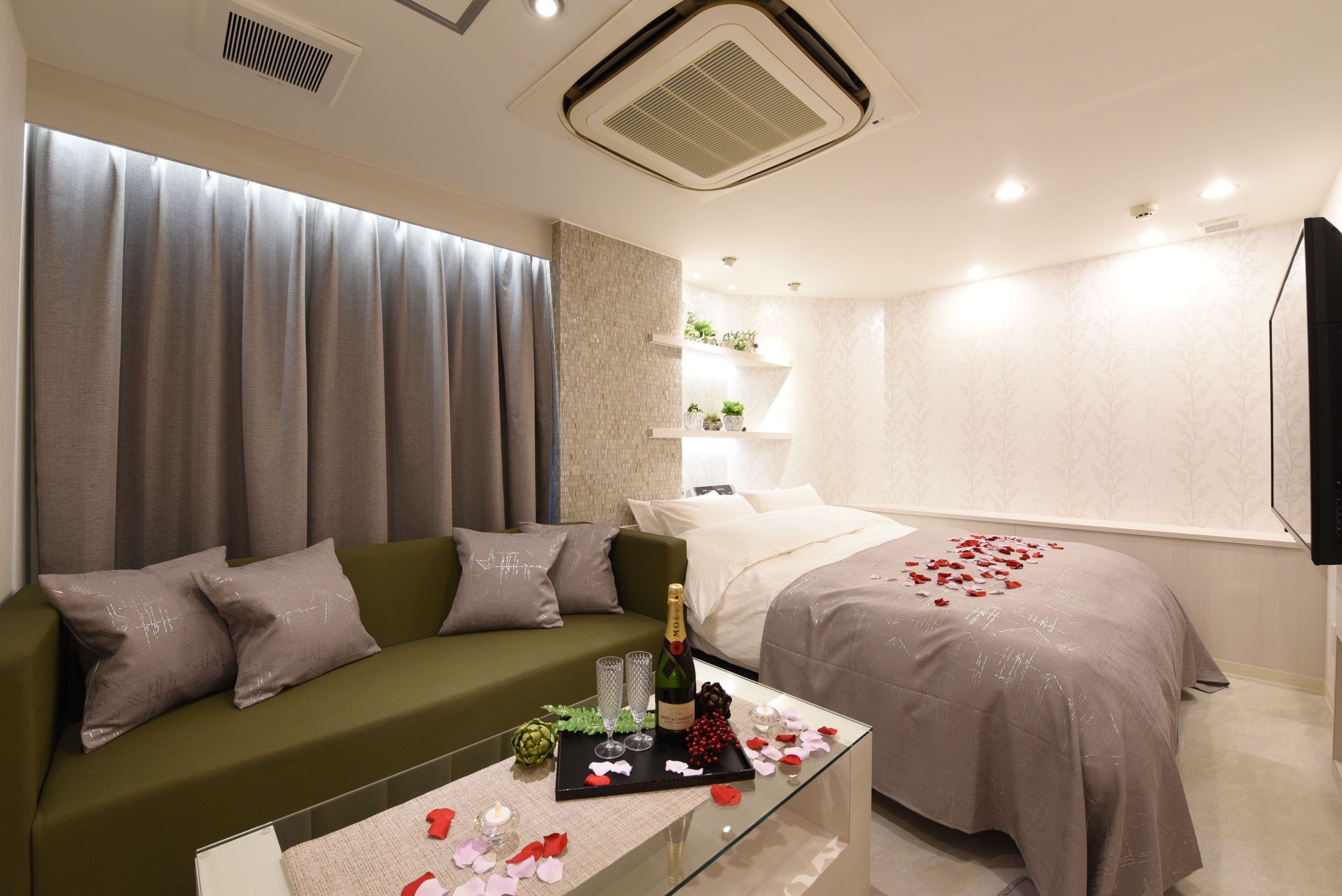 Una habitación de hotel moderna con un sofá verde, una mesa de centro de cristal con una botella y vasos, una cama con pétalos de rosa y un televisor en la pared.