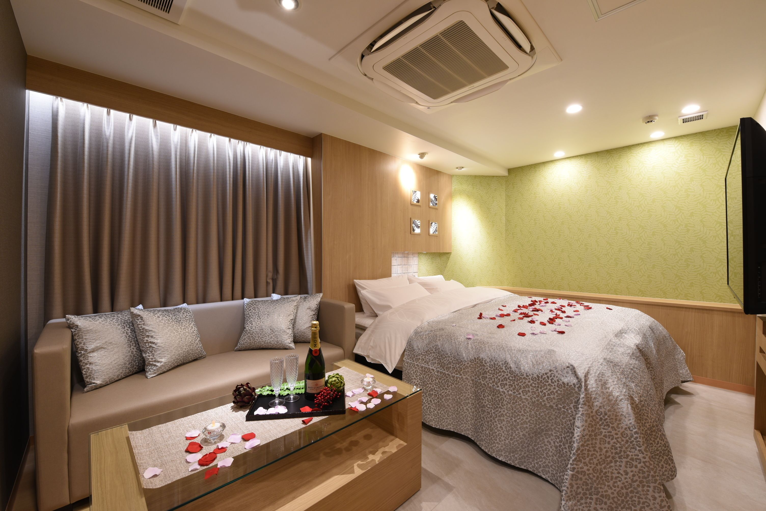 Una habitación de hotel con sofá, cama, una mesita de vidrio con pétalos de rosa y una botella, y pared verde.