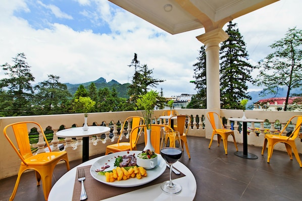 A terrace with yellow chairs and a table, a plate of food, and a glass of red wine.