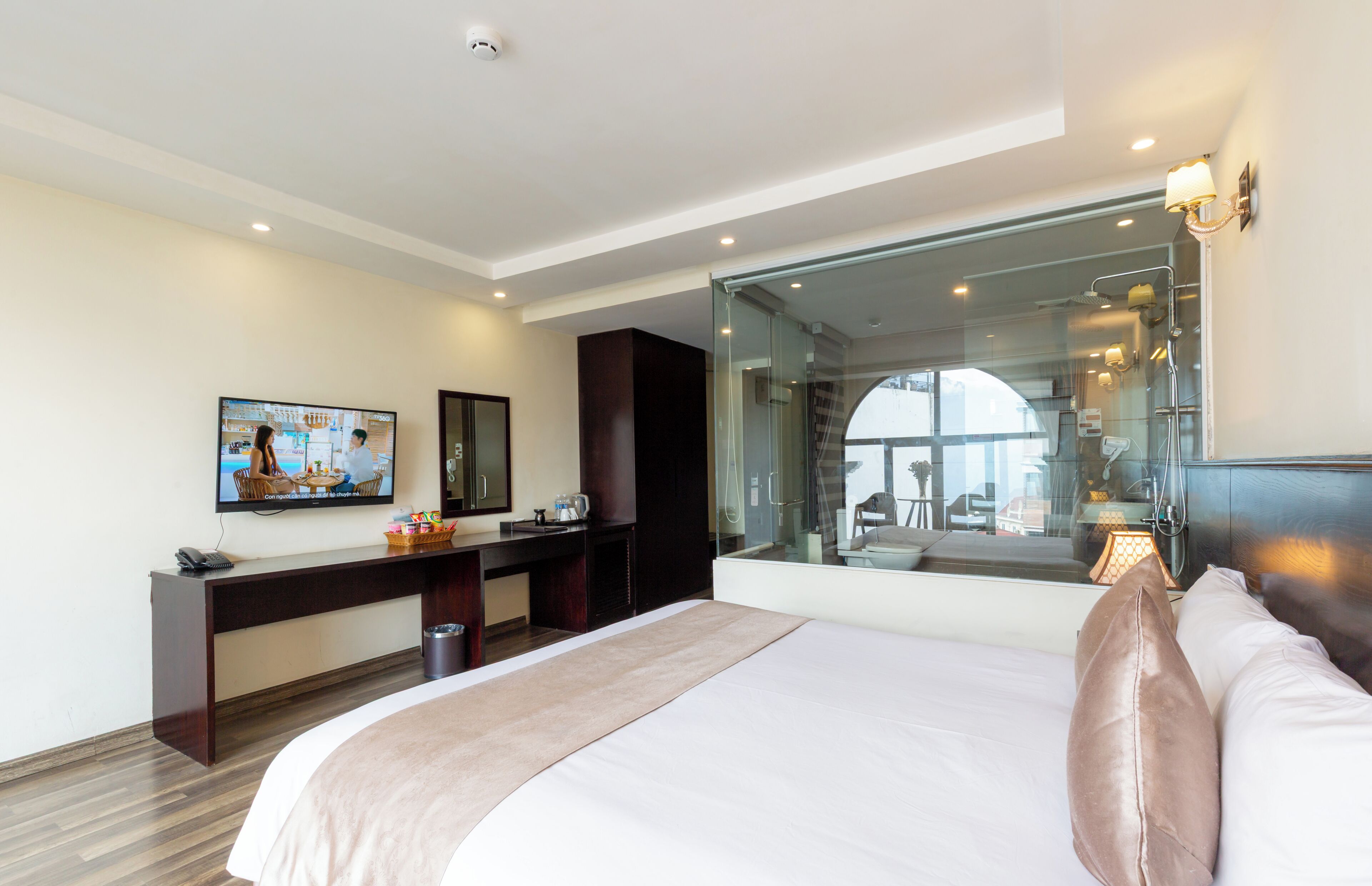 A modern hotel room with a large bed, a desk, a television, and a glass partition.