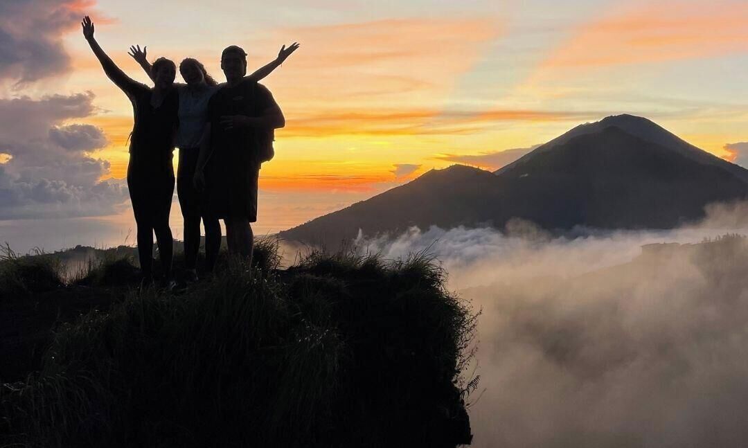 Three people standing on a grassy hill with their arms raised, overlooking a mountain and a body of water during sunset.