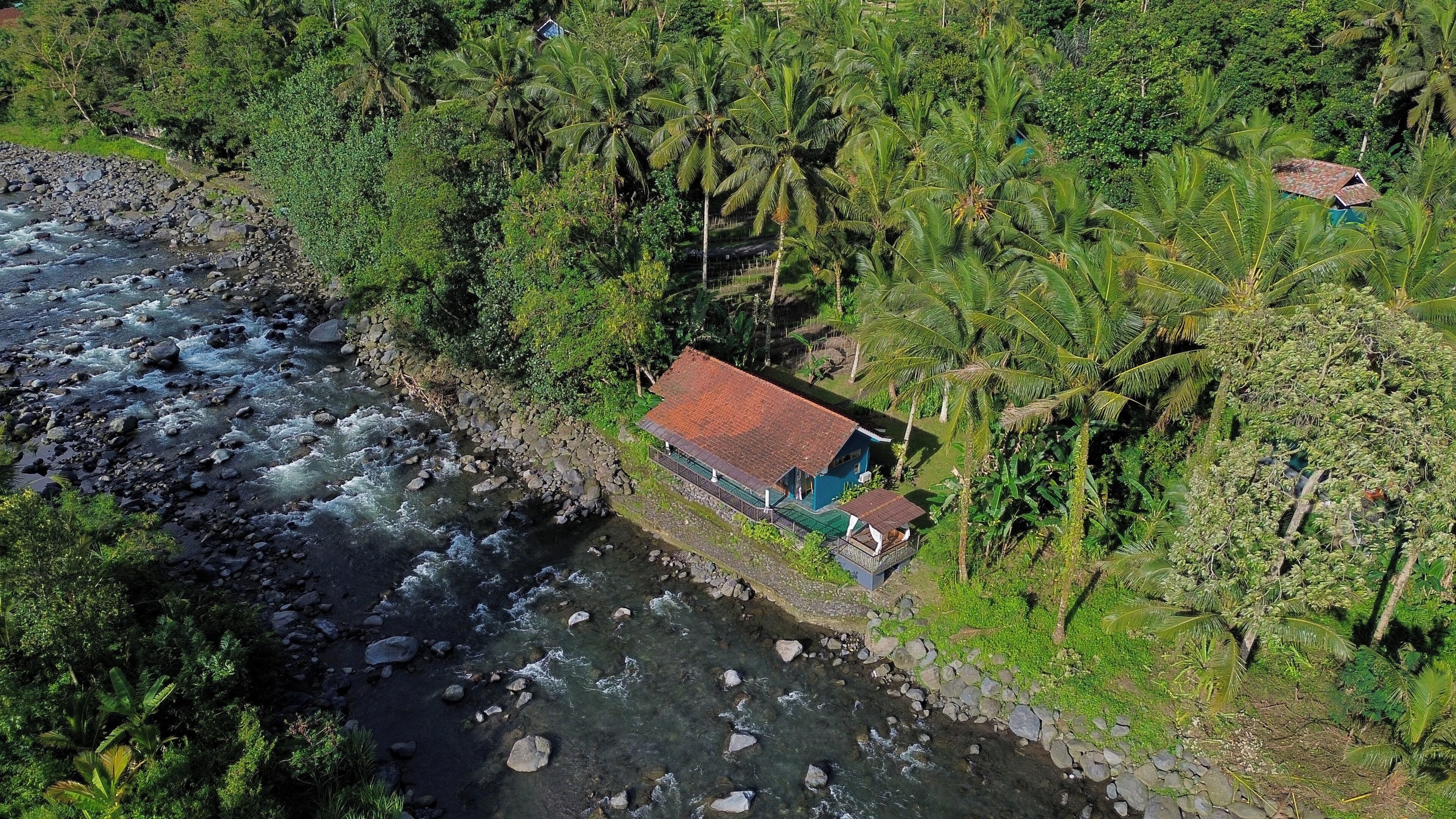 A house with a red roof, surrounded by dense tropical vegetation and a river.