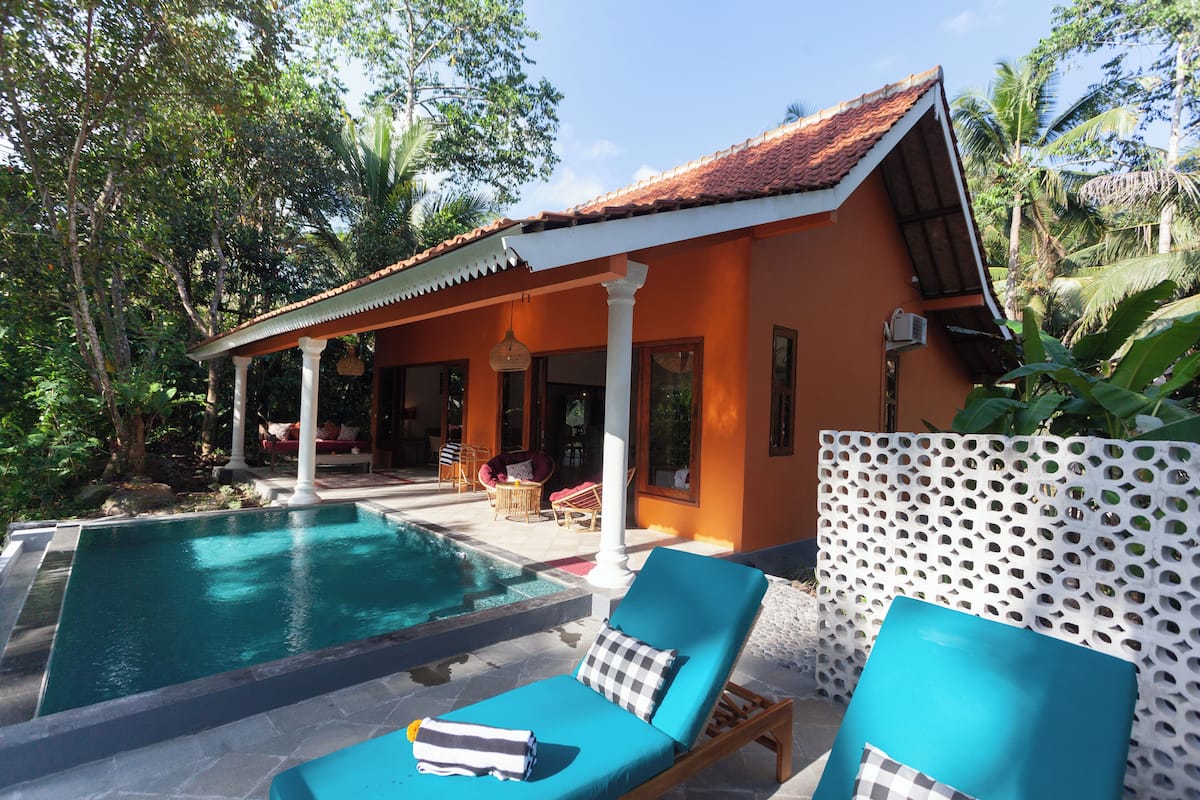 Un área junto a la piscina con sillones de descanso, un porche cubierto y vistas a un jardín tropical.