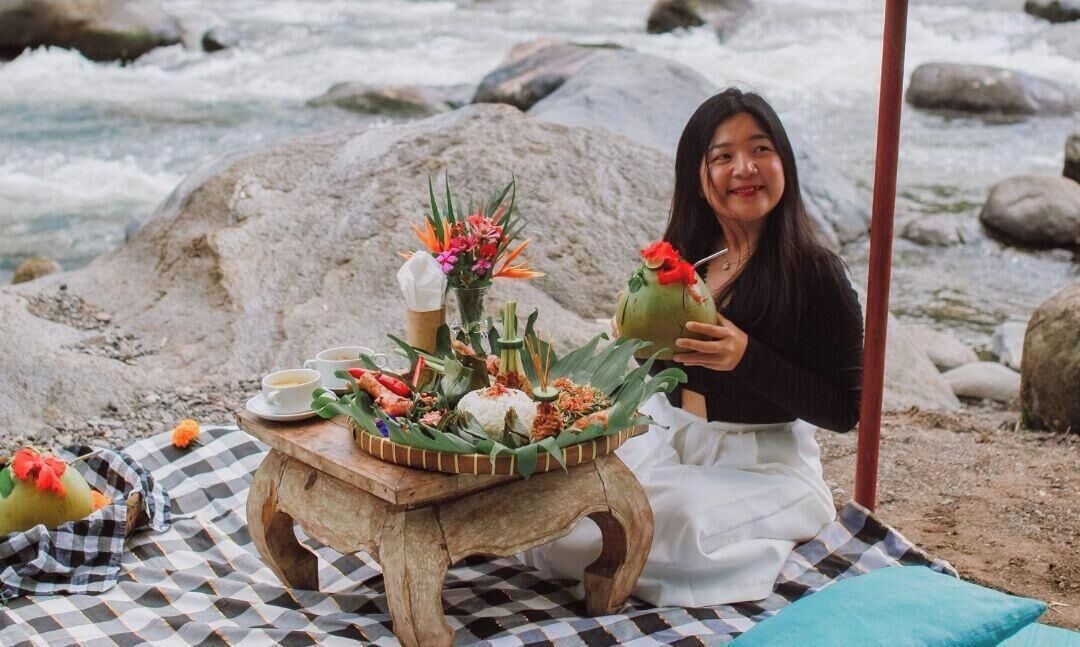Une personne assise sur une couverture à carreaux, devant une table dressée avec de la nourriture et des fleurs.