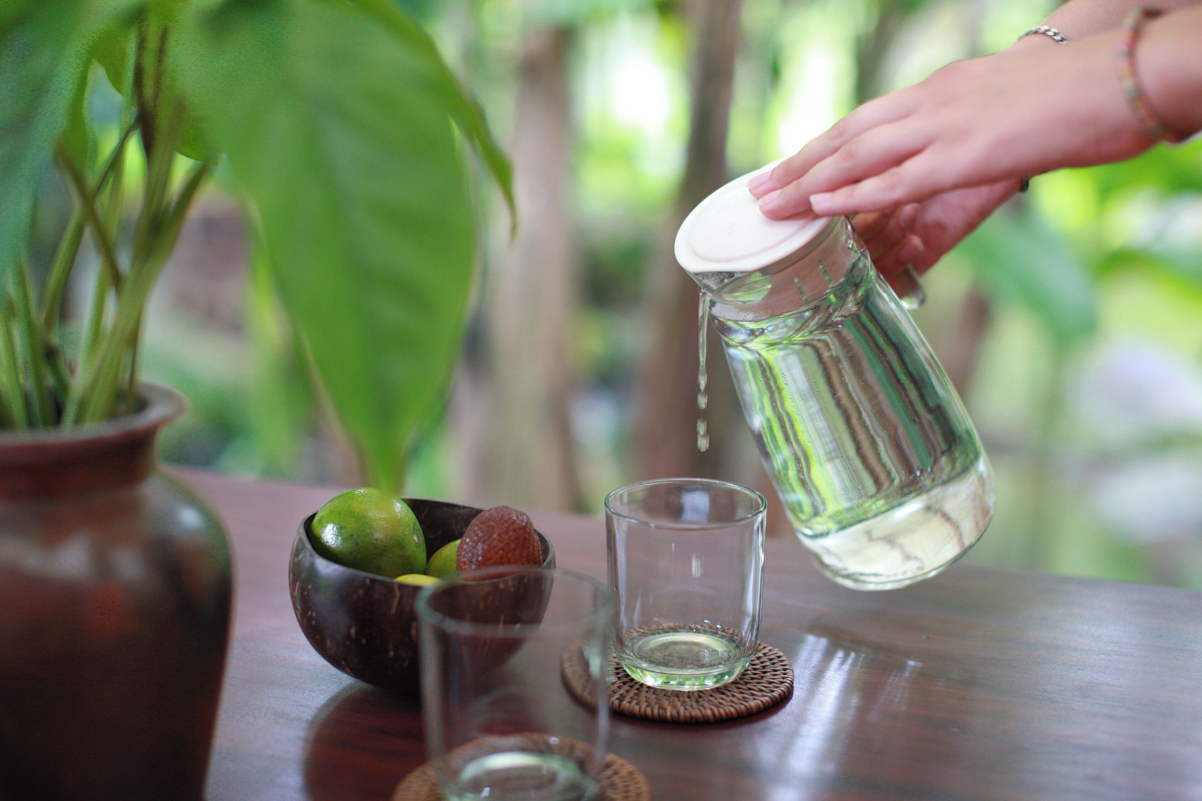 A hand pouring water from a pitcher into a glass on a wooden table with a bowl of fruit and a vase of green leaves.