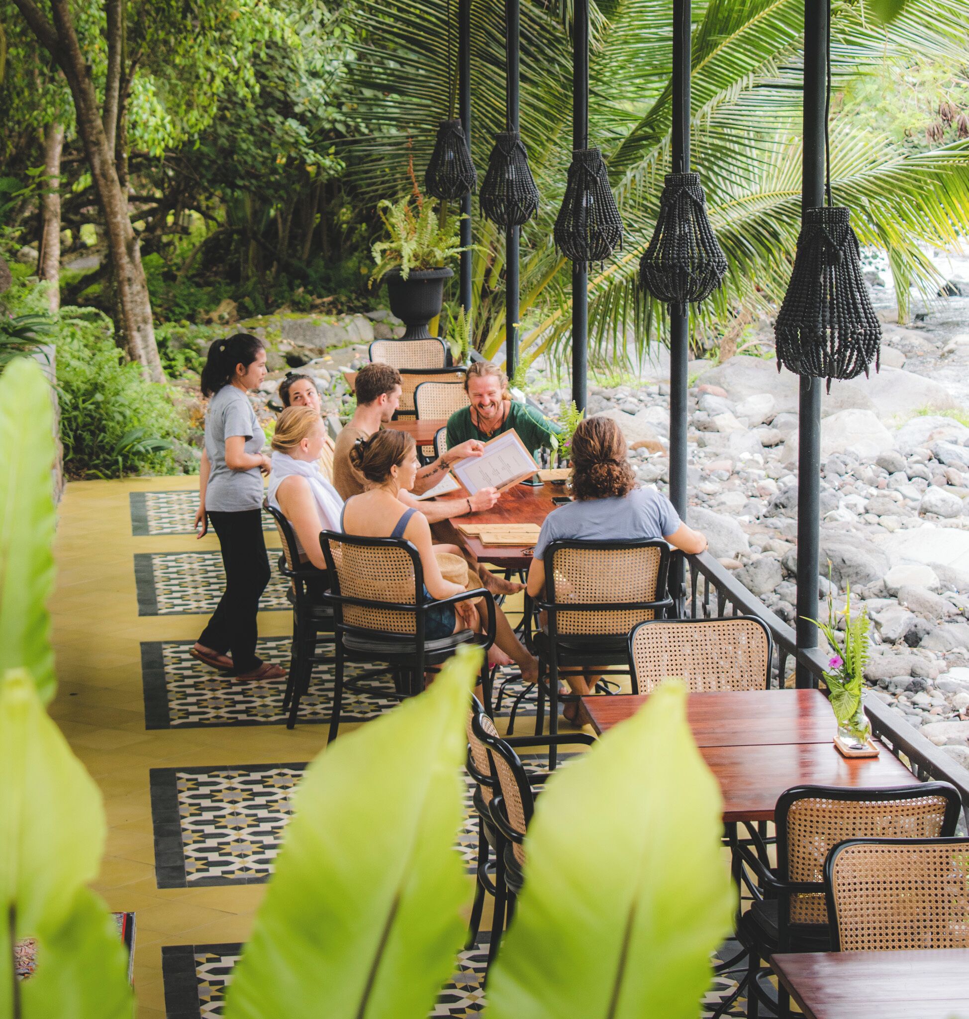 Un grupo de personas sentadas alrededor de una mesa en un entorno al aire libre, con un jardín tropical de fondo.