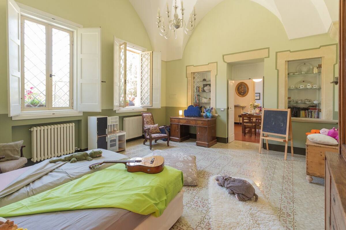 A spacious bedroom with a large bed, a desk, a chair, a guitar, and a small table with a teddy bear.
