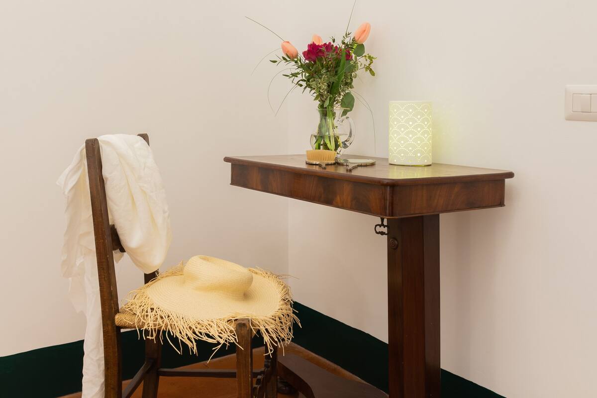 A room with a wooden chair, a small wooden table with a vase of flowers, and a framed painting on the wall.