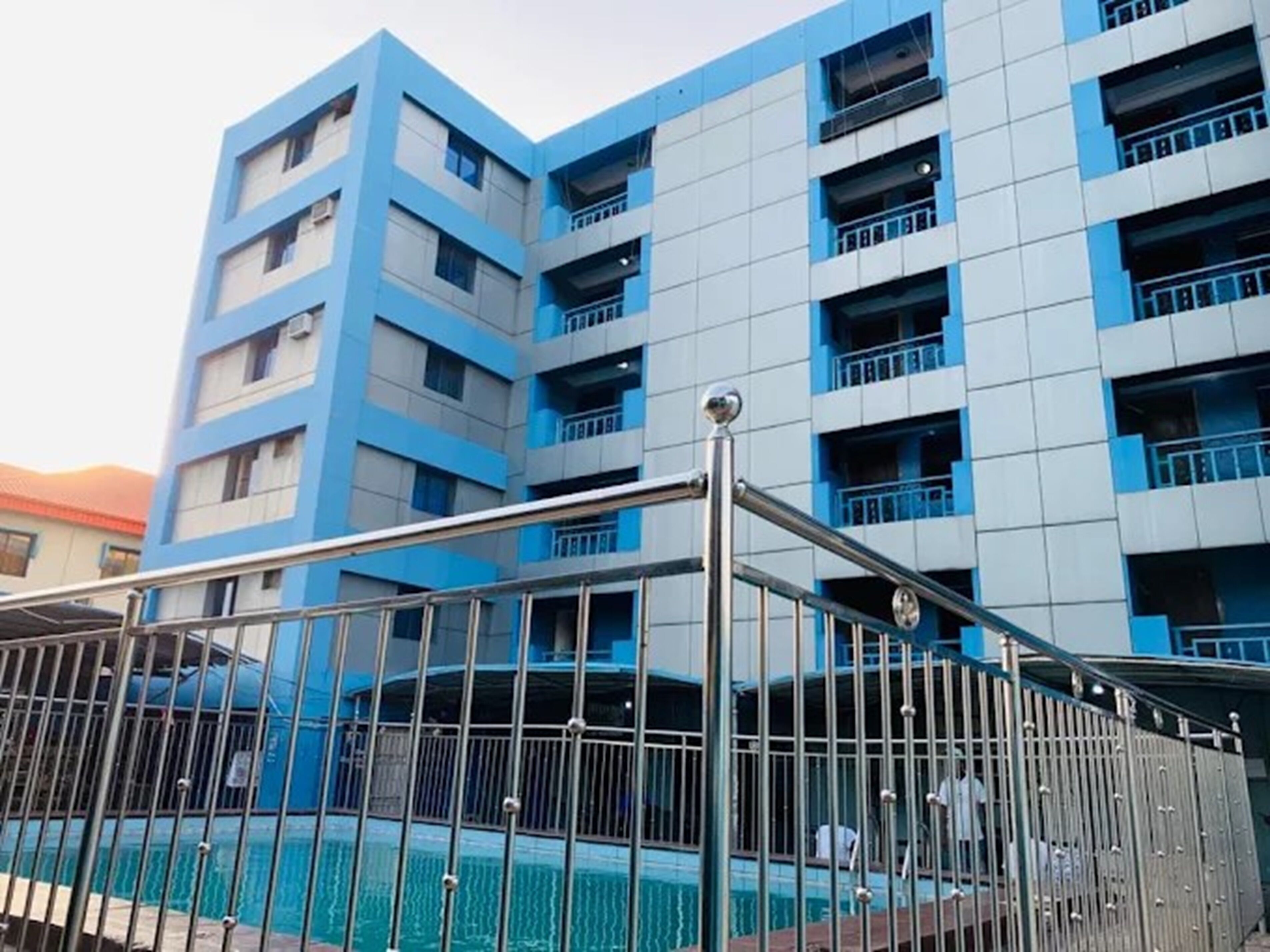 Un edificio de varios pisos con fachada azul y blanca, una piscina y una cerca de metal.