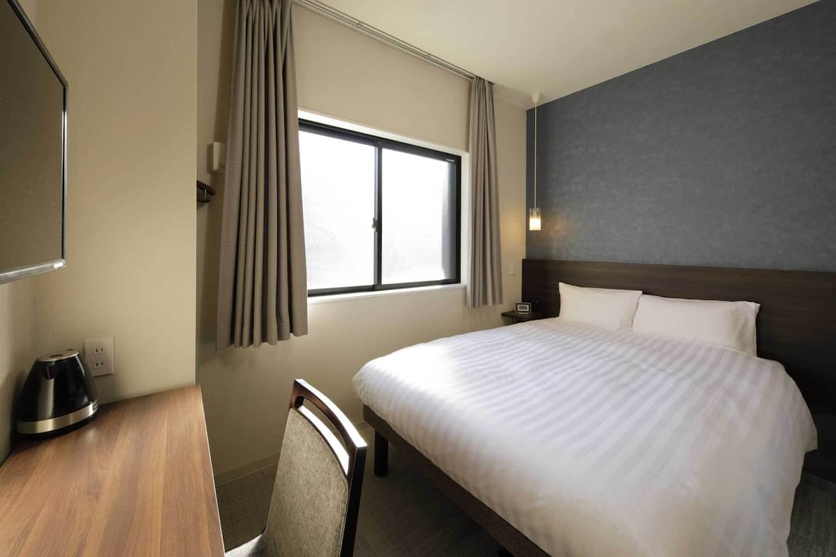 A hotel room with a large bed, a desk with a kettle, a chair, a window with curtains, and a wall-mounted light fixture.