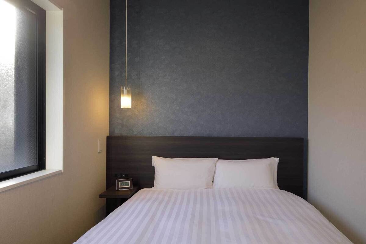 A hotel room with a bed, a nightstand, a wall clock, and a window.