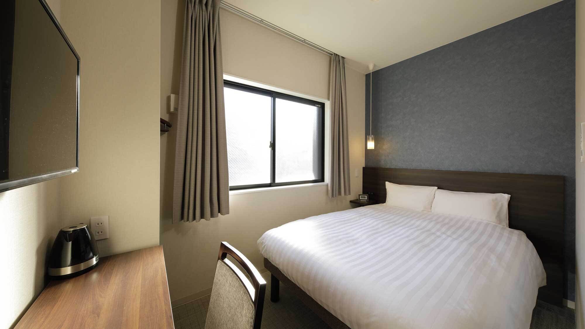 A hotel room with a bed, a desk with a kettle, a chair, a window with curtains, and a wall-mounted light.