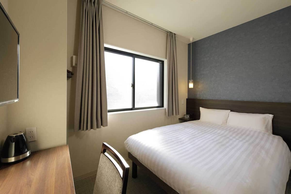 A hotel room with a bed, a desk with a kettle, a chair, a window with curtains, and a wall-mounted light.