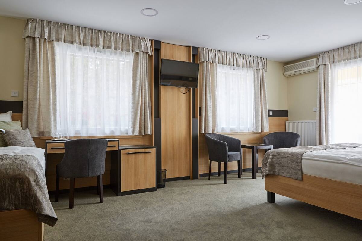 A hotel room with two beds, a desk with a chair, a TV, and large windows with curtains.