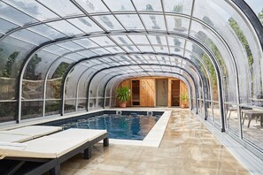 Una piscina cubierta con vidrio, sillas de descanso, piso de madera y plantas en macetas.