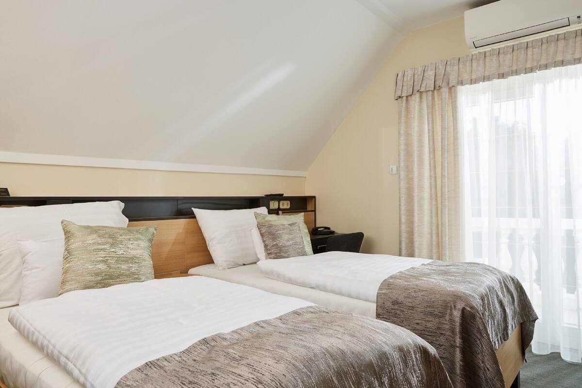 A hotel room with two beds, a wooden headboard, a nightstand, and a window with sheer curtains.