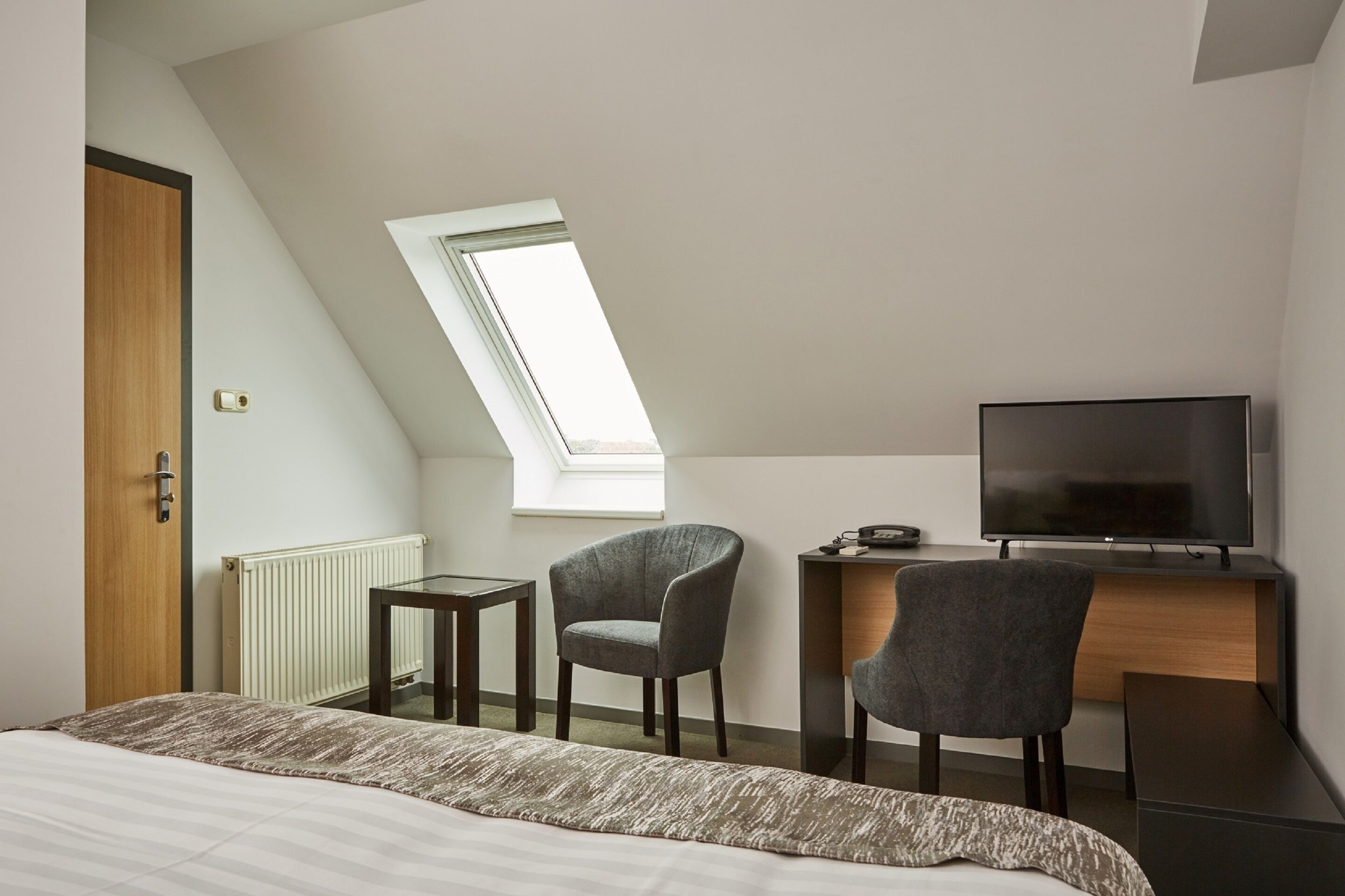 A modern hotel room with a large bed, a desk with a TV, two chairs, a small table, and a skylight.