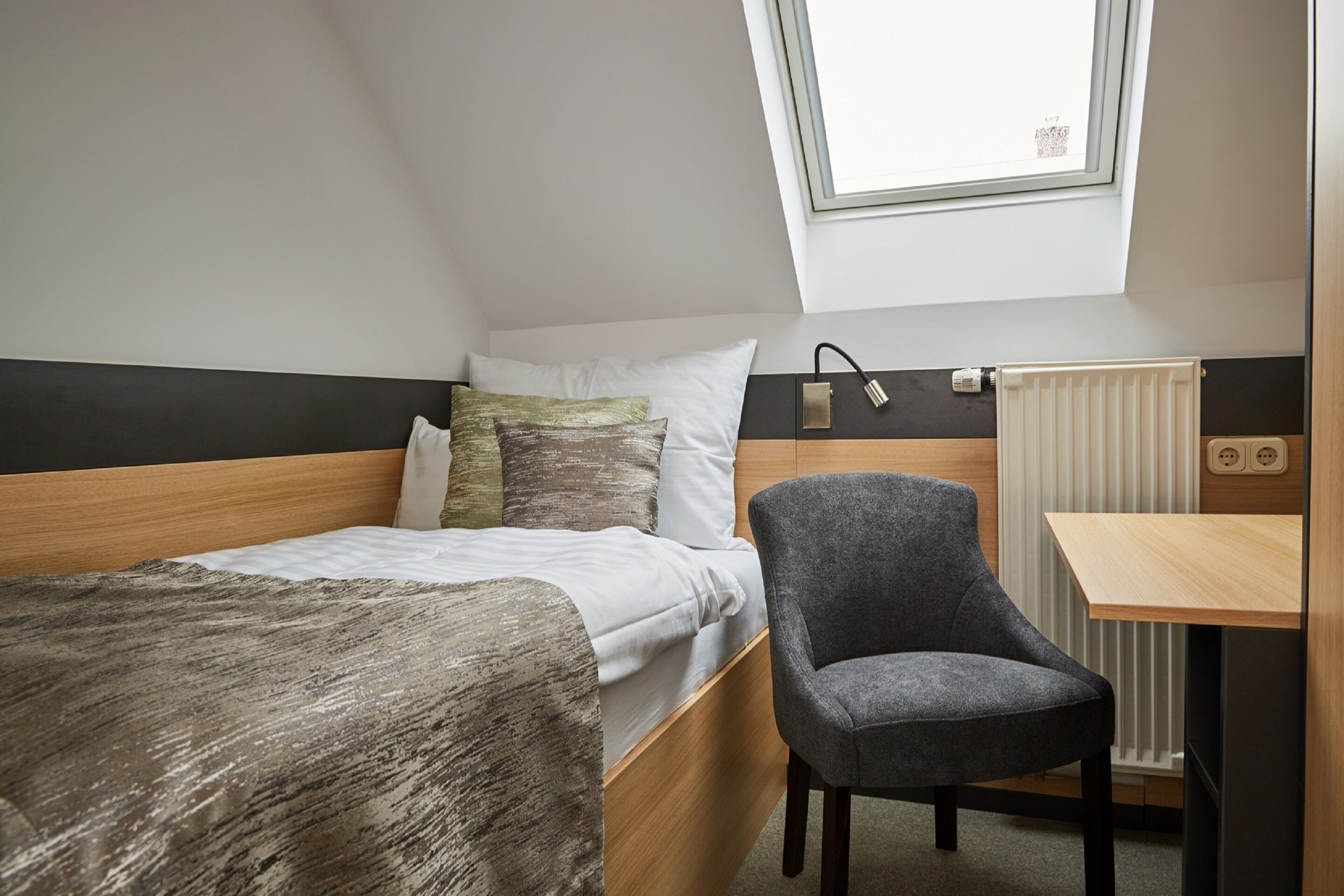 A compact bedroom with a single bed, a gray chair, a small table, and a skylight.