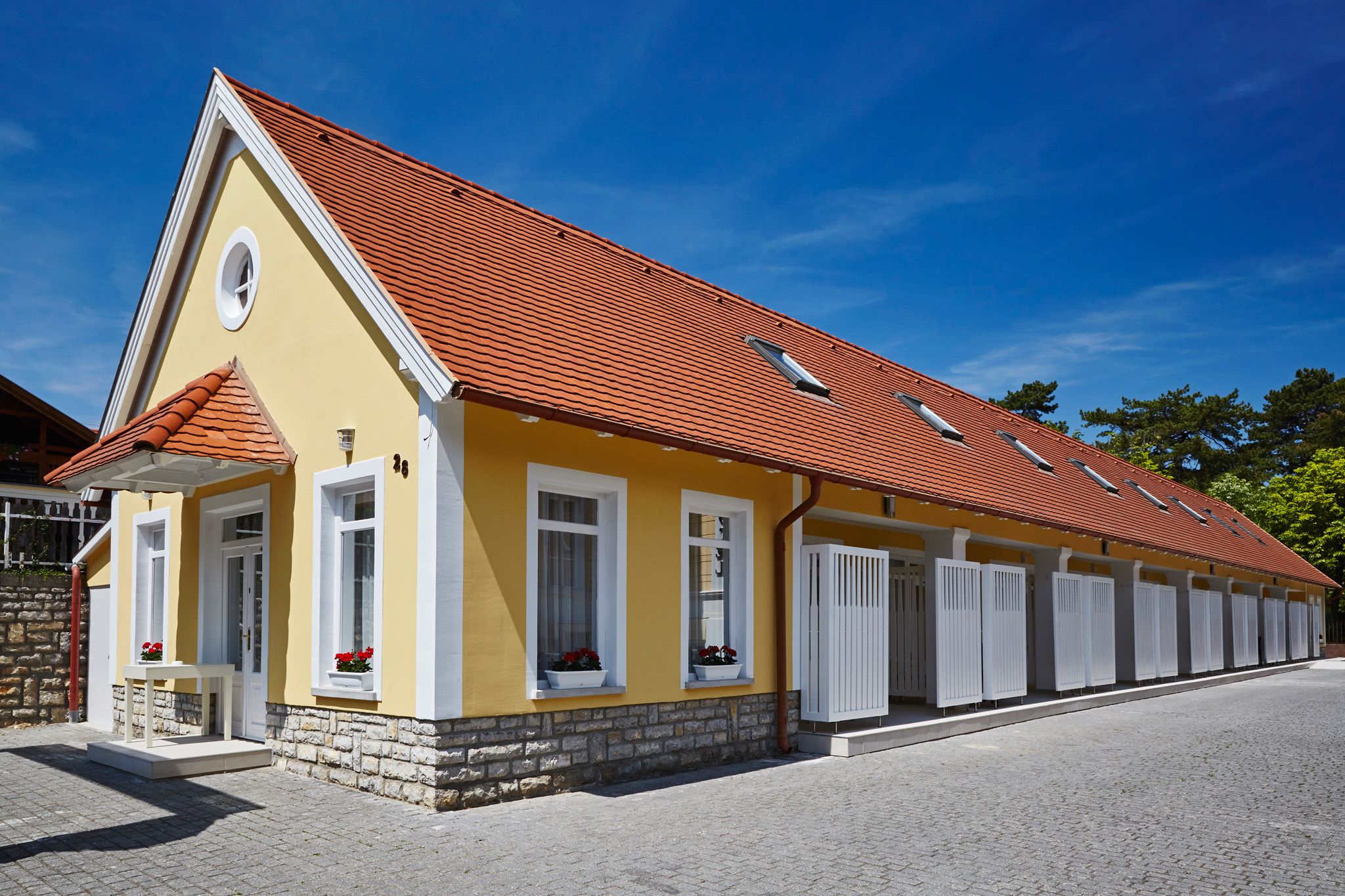 A yellow building with a red roof, white windows, and a stone base.