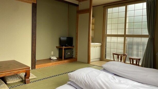 A traditional Japanese-style room with tatami flooring, a low wooden bed, a television on a cabinet, and a sliding door leading to a bathroom. - Oyado Uchidaya (Nozawaonsen)