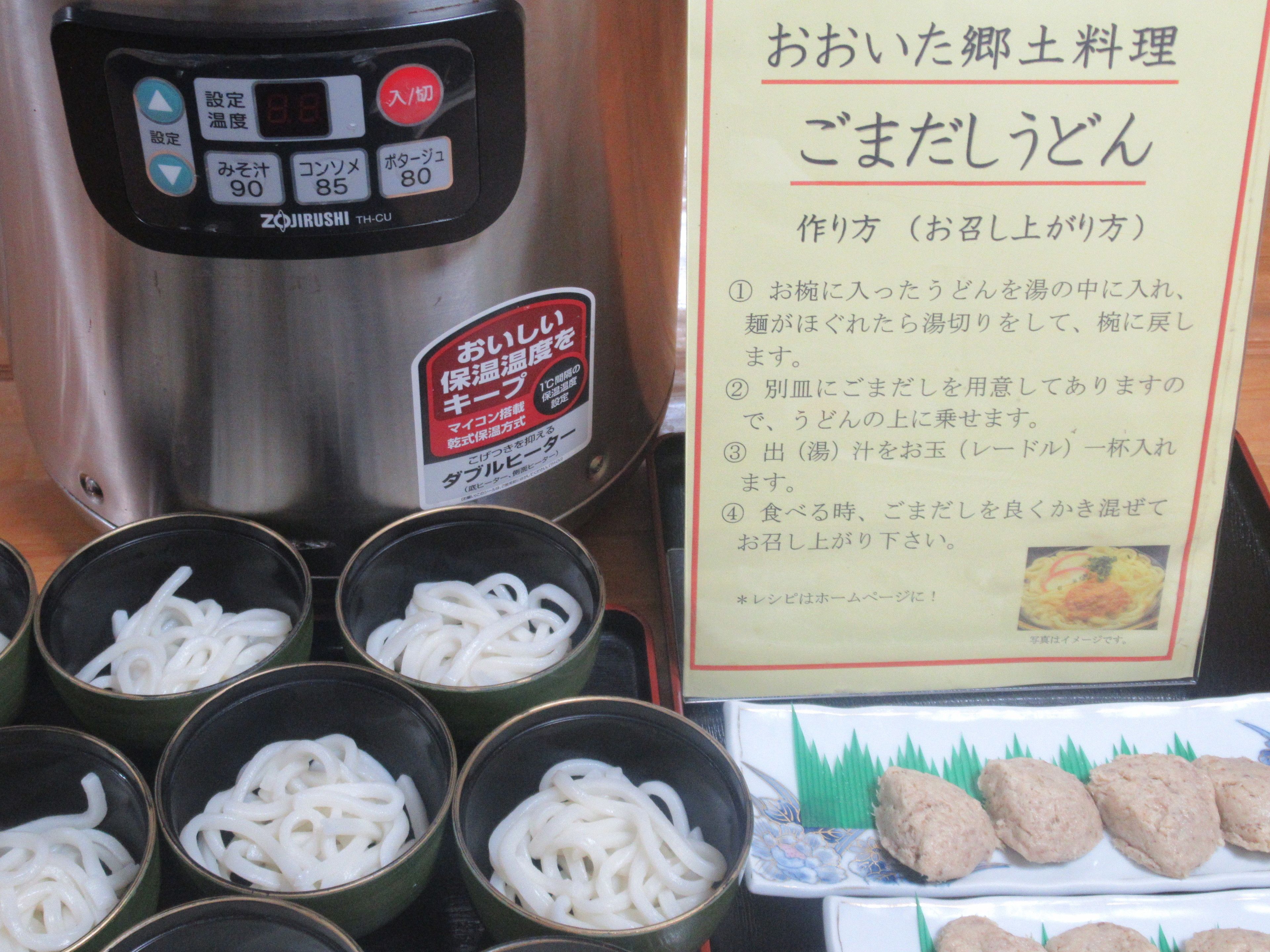 A kitchen area with a rice cooker, bowls of cooked noodles, and a menu board.
