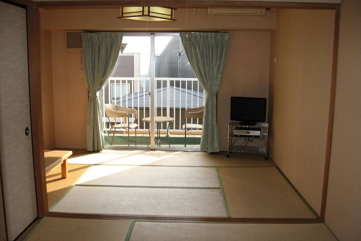 Une pièce de style japonais traditionnel, avec un sol recouvert de tatamis, une table basse, deux chaises, un téléviseur posé sur un meuble et un balcon offrant une vue.