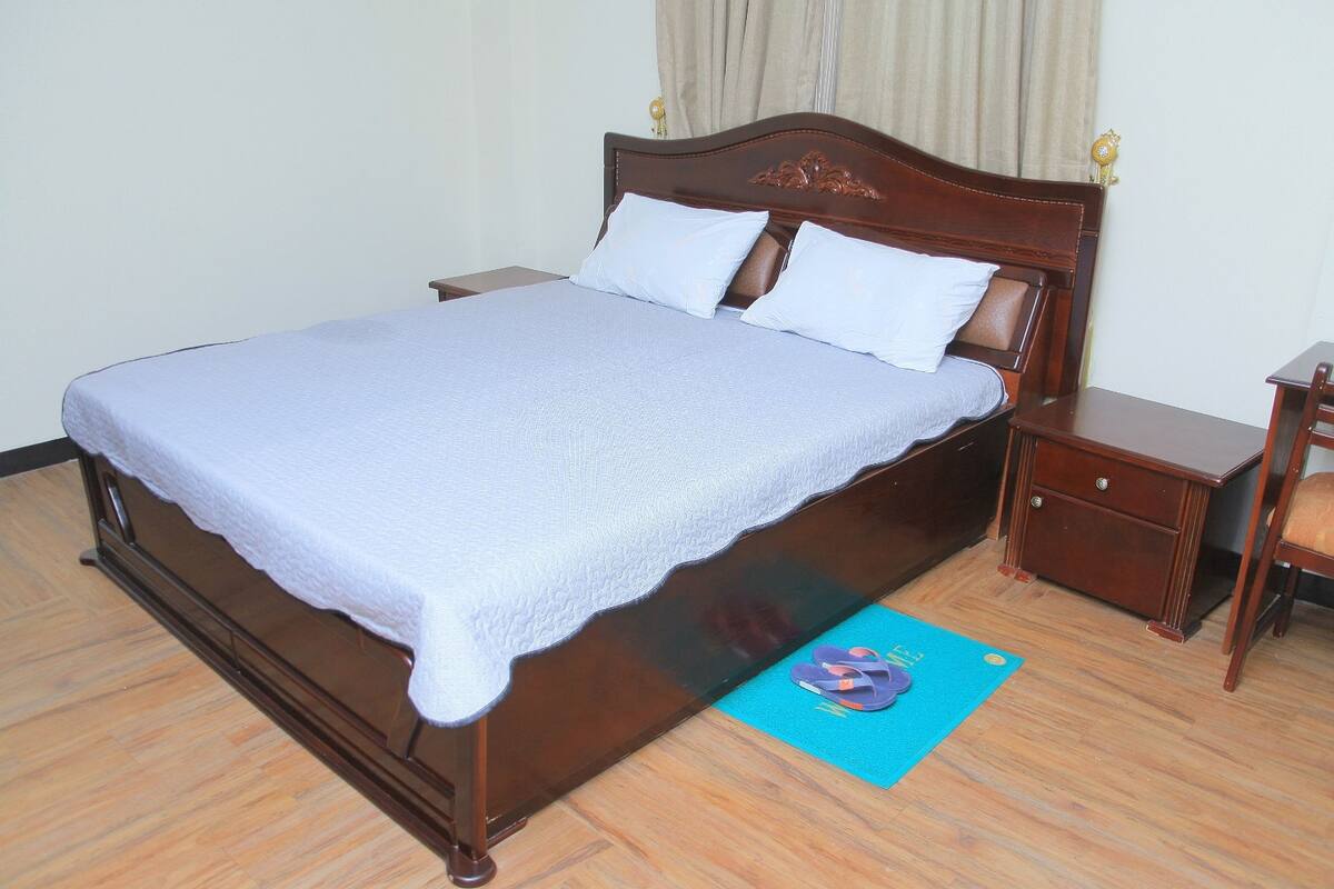 A neatly made bed with a white bedspread and two pillows, a wooden nightstand with a drawer, and a blue mat on the floor.