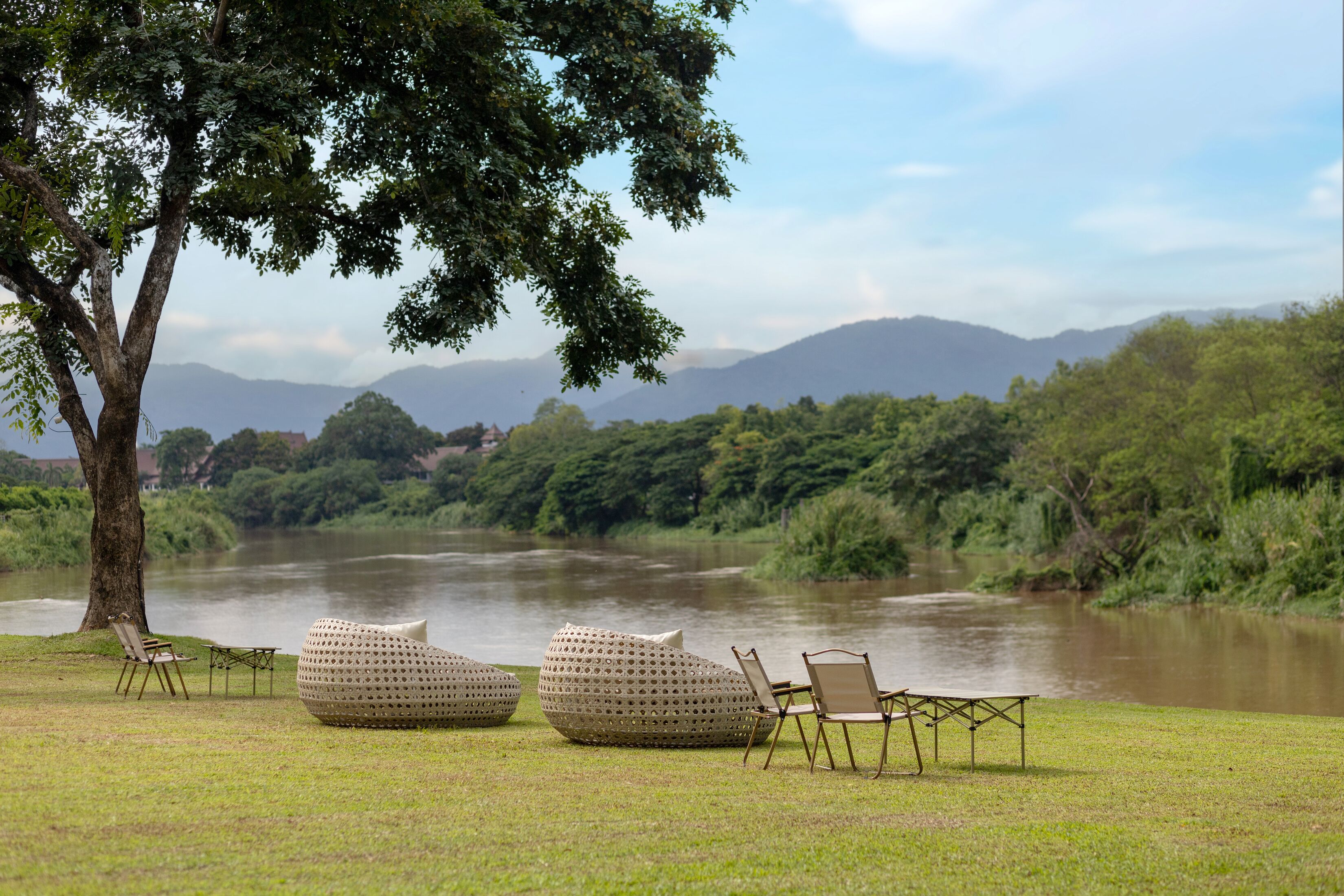 A serene riverside setting with seating arrangements and a large tree providing shade.
