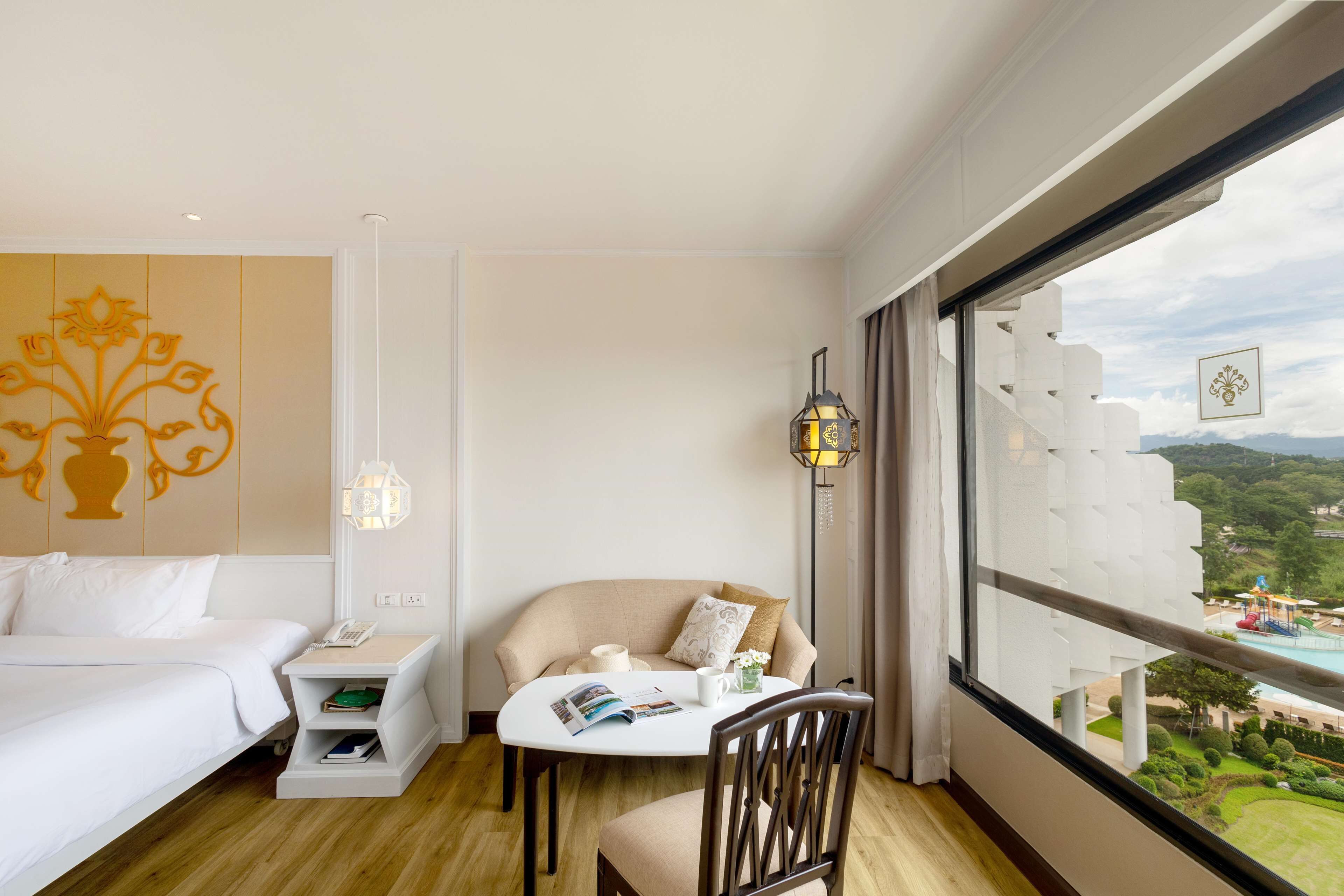 A hotel room with a bed, a small table with a magazine, a chair, a nightstand, and a window with a view of a pool and landscape.