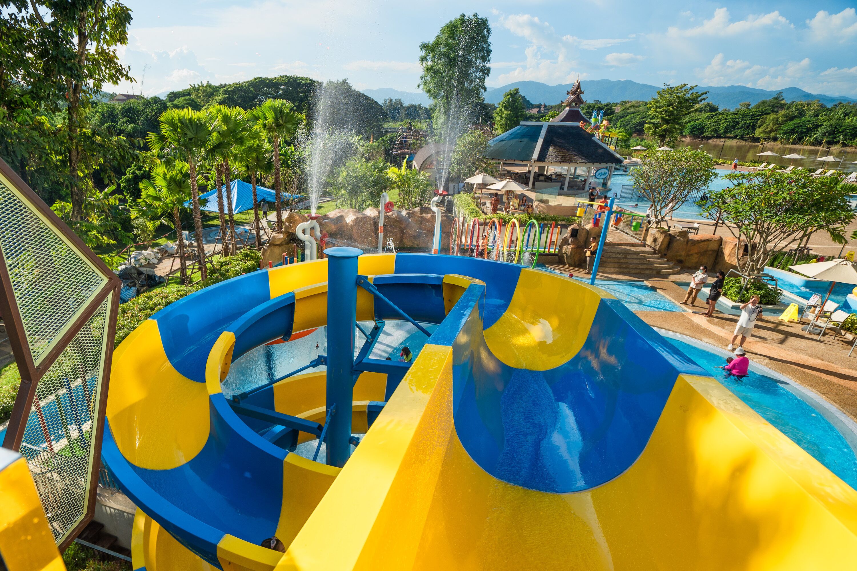 Un parco acquatico con uno scivolo grande, blu e giallo, una zona piscina e una vista sulle montagne sullo sfondo.