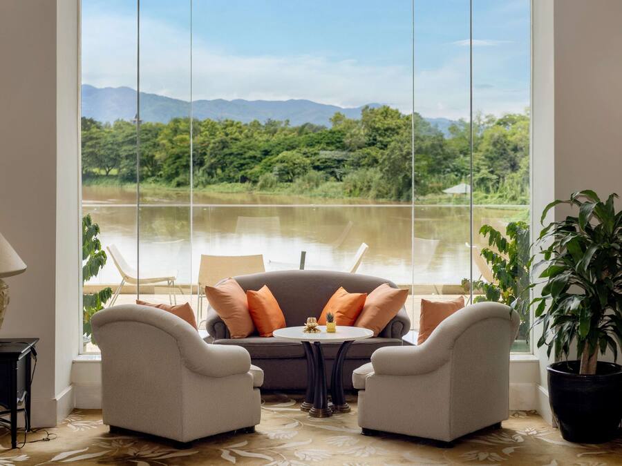 Habitación con un ventanal grande que ofrece vistas a un lago y montañas. Hay un sofá con dos sillones, una mesita y una lámpara.