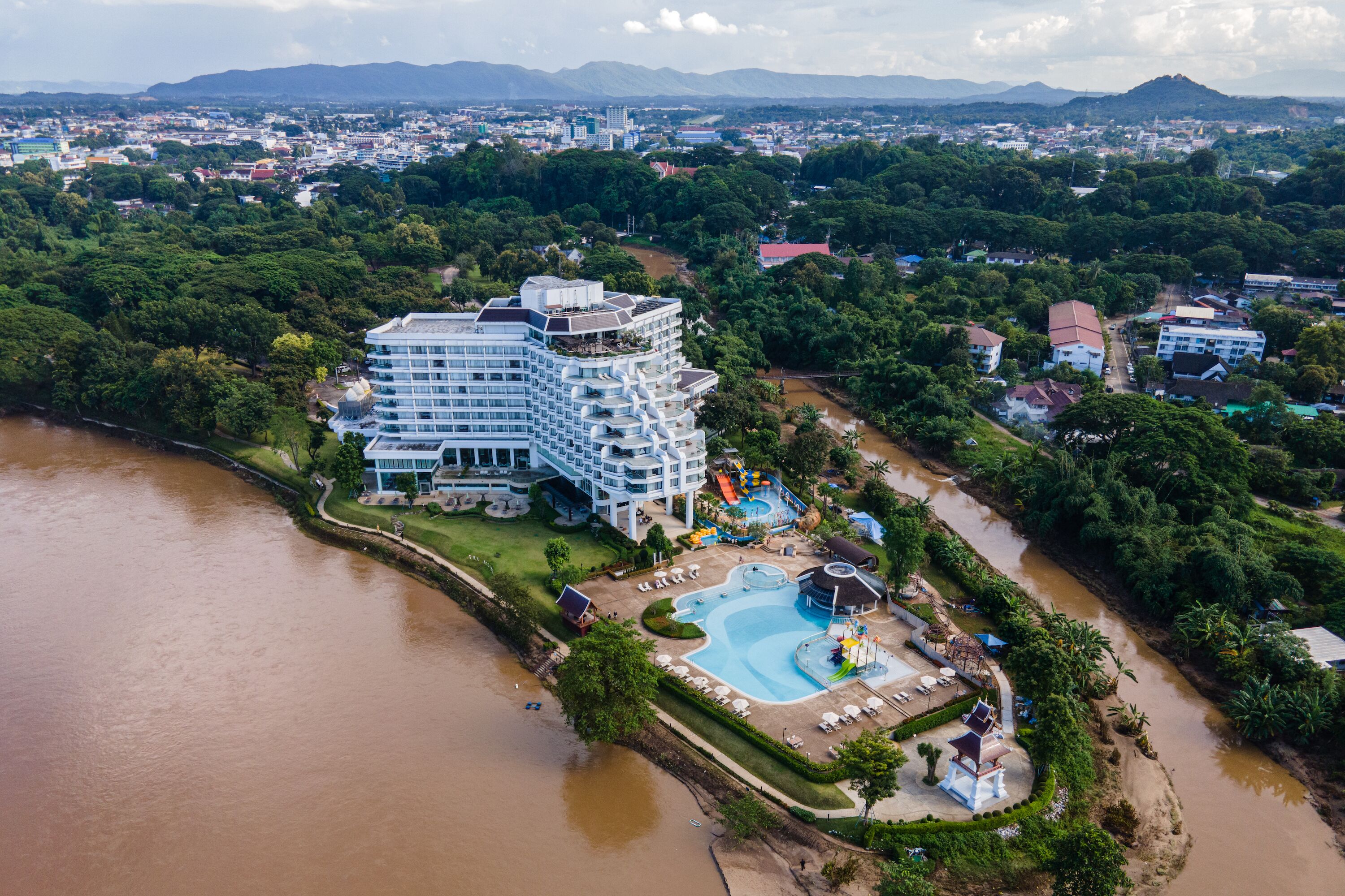 An aerial view of a resort with a large pool, a water slide, and a view of the surrounding city and mountains.