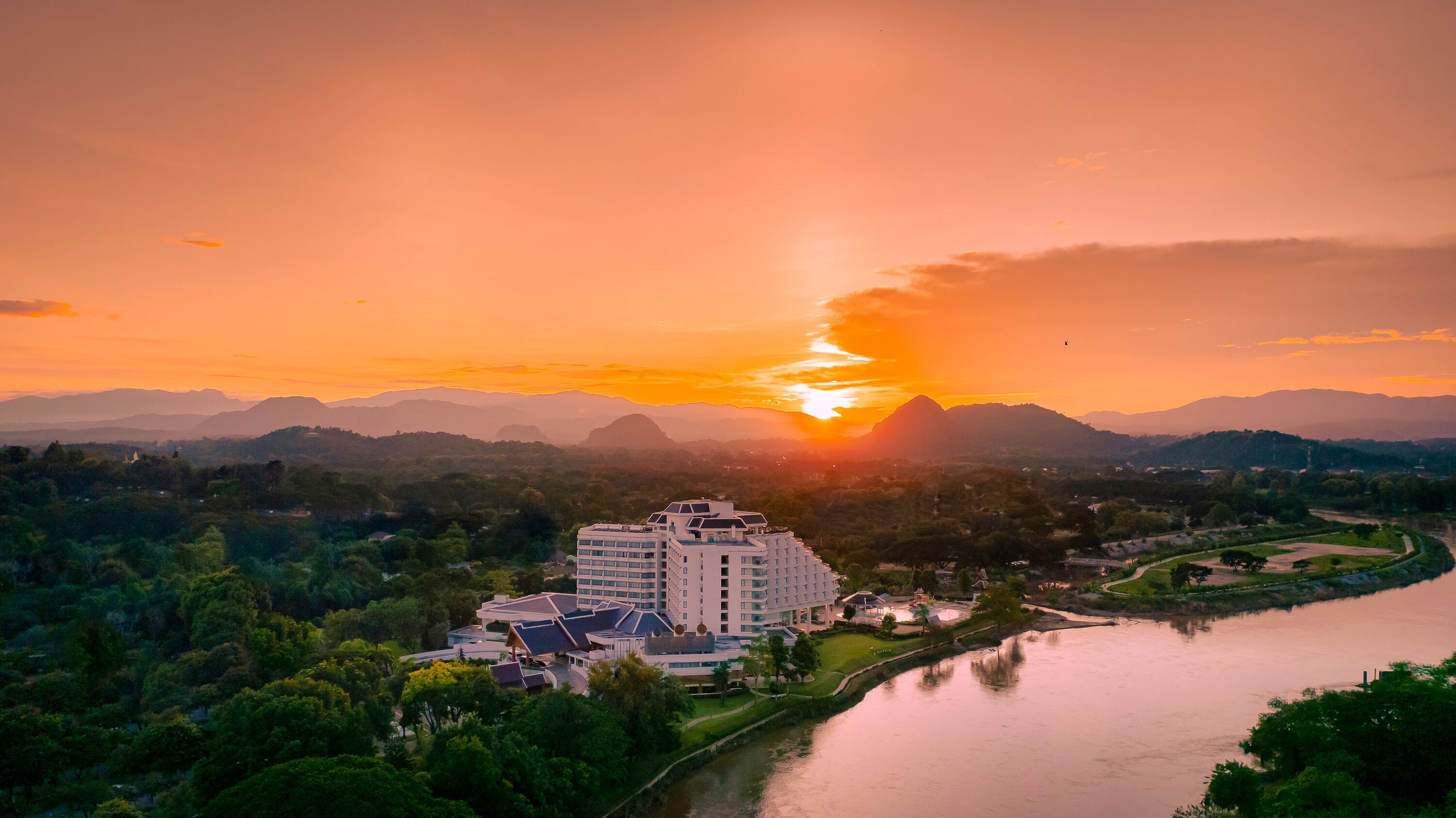 A modern hotel with multiple floors, surrounded by lush greenery and a river.