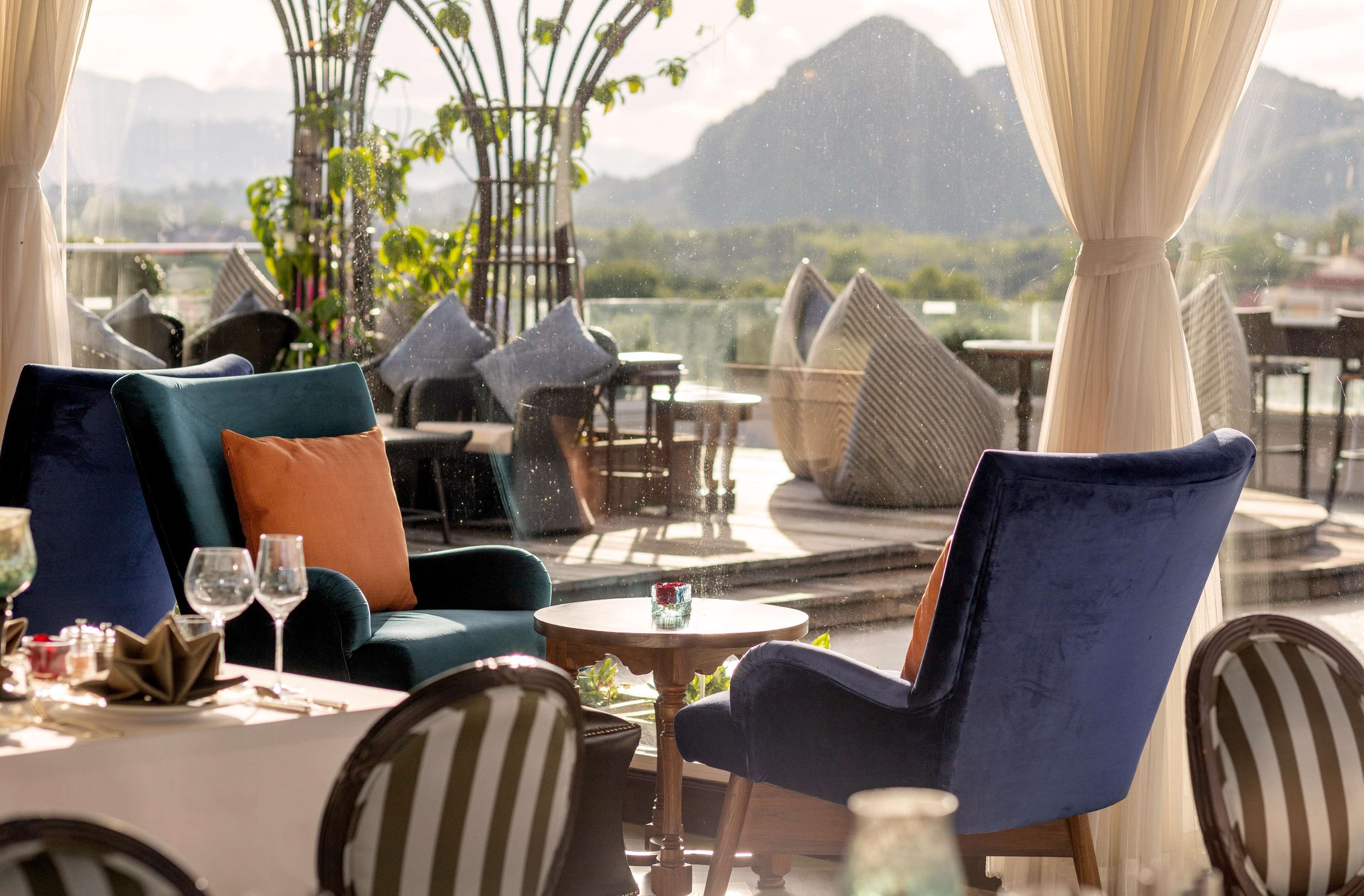 A restaurant with outdoor seating, including cushioned chairs and tables, set against a scenic mountain backdrop.