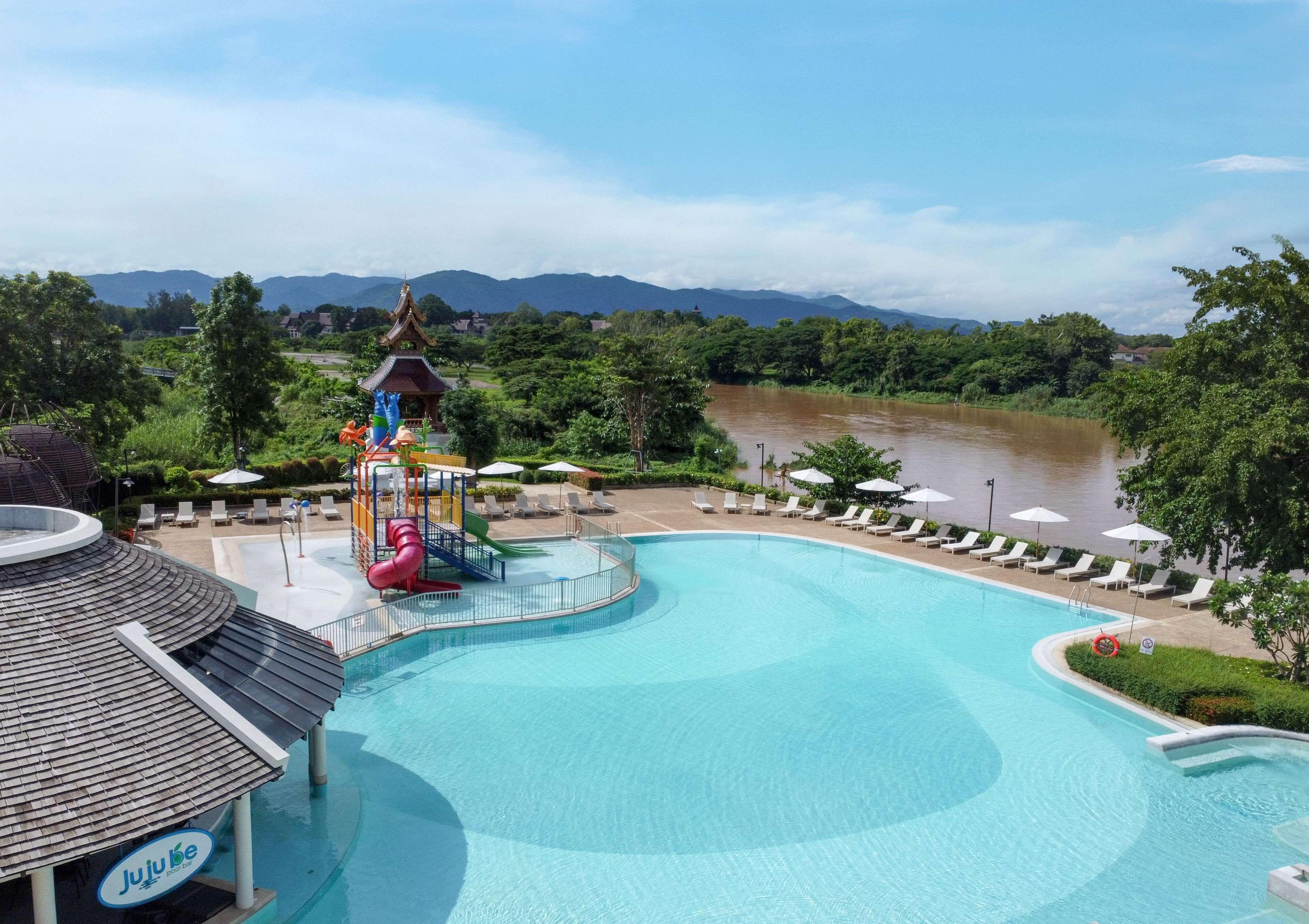 A swimming pool with a water slide and a pavilion, surrounded by lounge chairs and umbrellas, with a river and mountains in the background.
