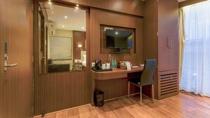 A hotel room with a wooden desk, a television mounted above it, a chair, a trash bin, and a large window with curtains. - Hotel Sawood International (Kolkata)