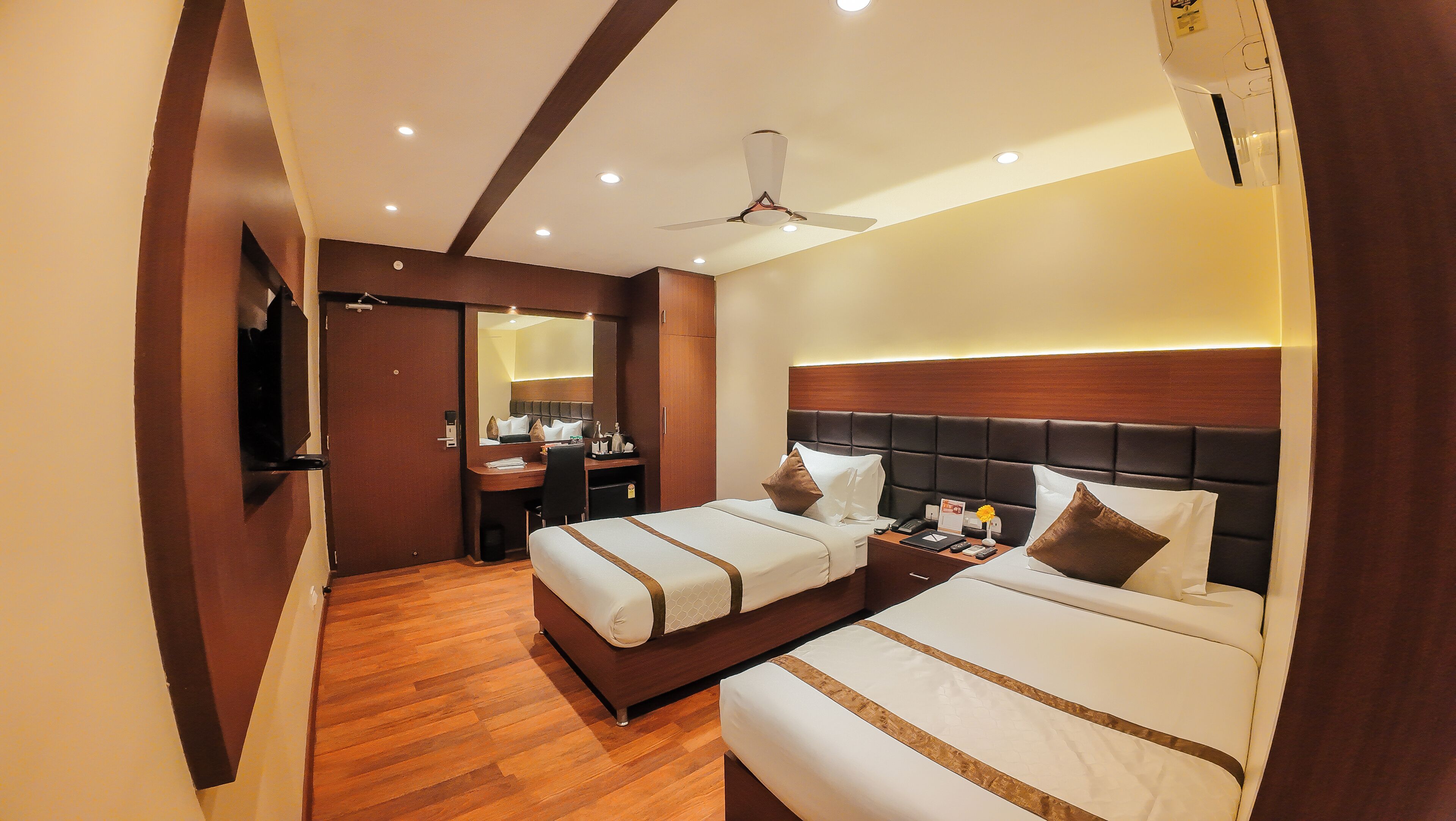 A hotel room with two beds, a desk, a mirror, and a ceiling fan.