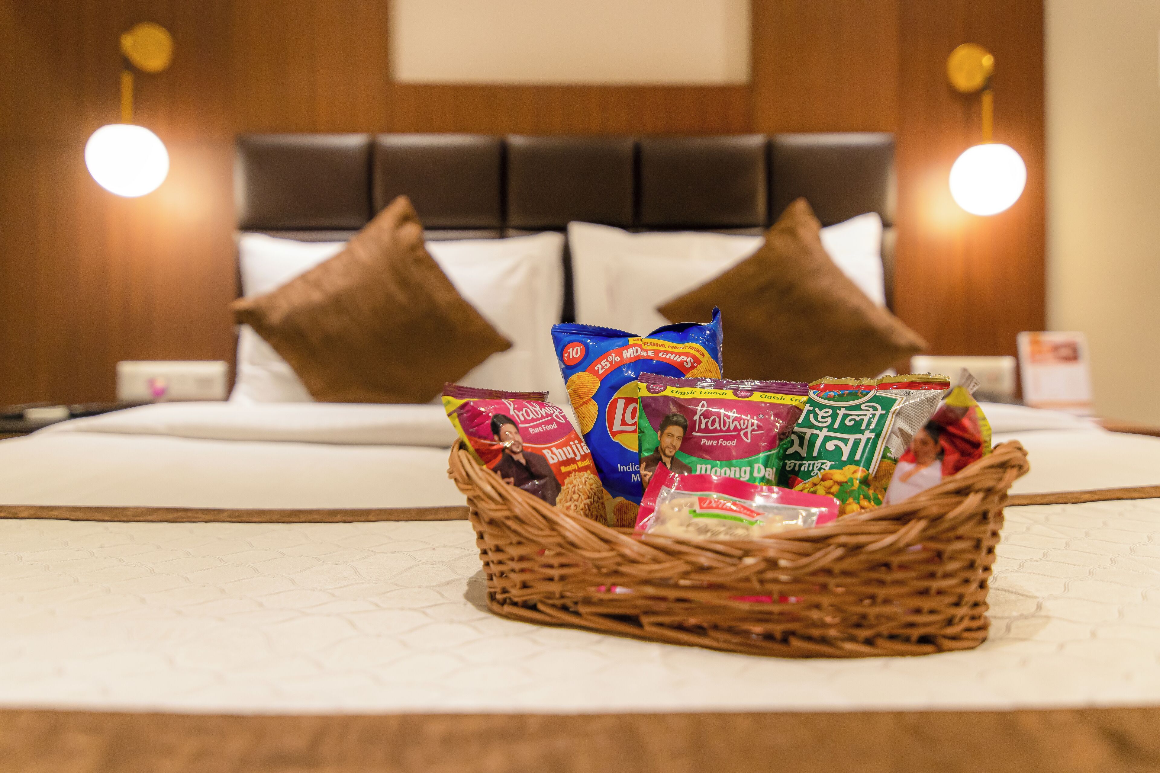 A hotel room with a neatly made bed, a bedside table with a lamp, and a wicker basket filled with snacks on the bed.