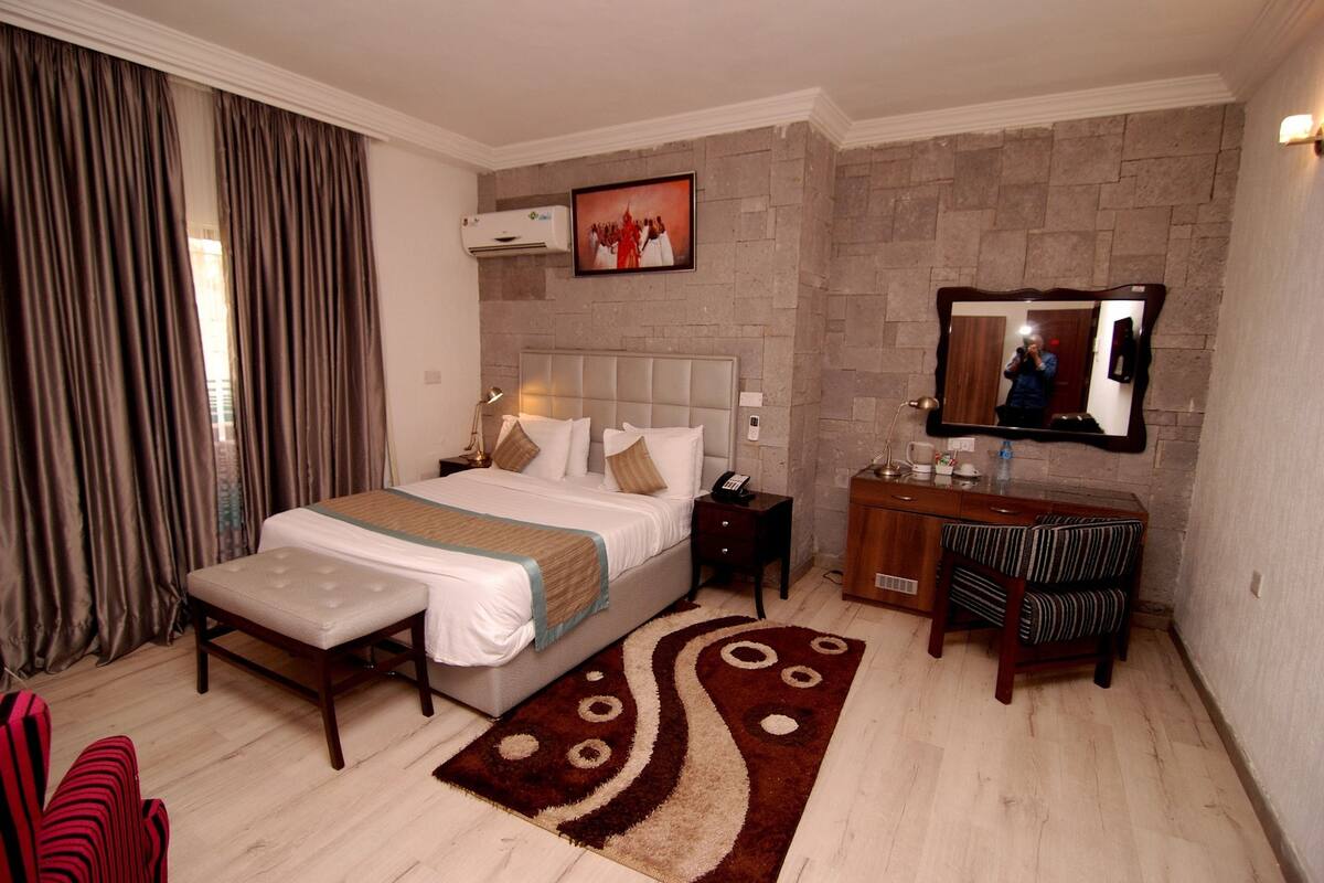 Ein Hotelzimmer mit Bett, Nachttischen, Schreibtisch, Stuhl, Spiegel und einem an der Wand befestigten Fernseher.