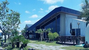 A modern building with a flat roof and large windows, surrounded by a stone-paved courtyard and palm trees. - Jeju Platinum Caravan (Jeju City)