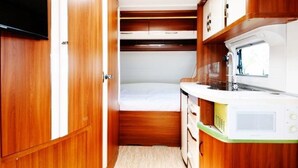 A compact kitchenette with wooden cabinets, a microwave, and a small bed. - Jeju Platinum Caravan (Jeju City)