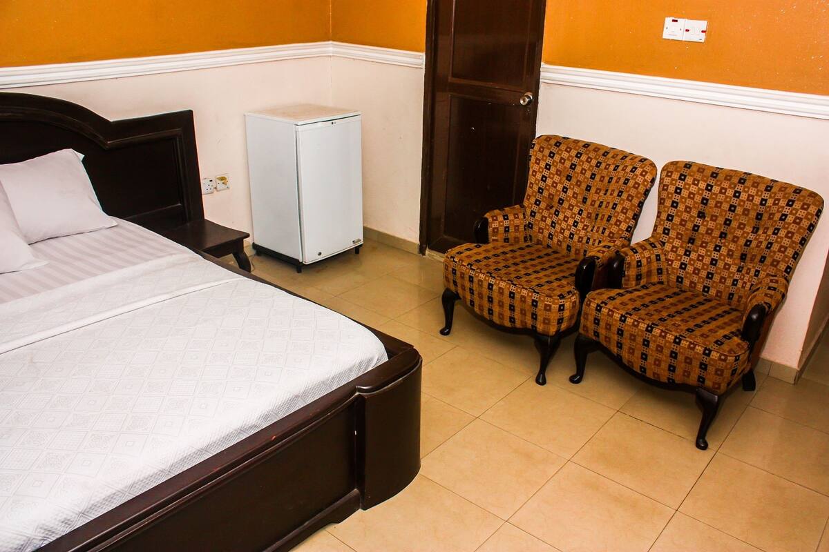 A bedroom with a bed, a small white refrigerator, and two patterned armchairs.