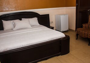 A neatly made bed with a dark headboard, a small bedside table, a white refrigerator, and a patterned armchair.