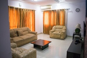Una sala de estar con un sofá, un sillón y una mesita de centro. Hay un aire acondicionado en la pared y un reloj también en la pared.