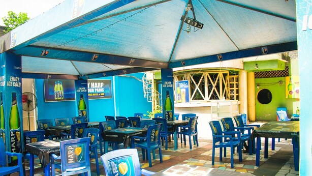 An outdoor seating area with blue chairs and tables, some covered by umbrellas, and walls adorned with advertisements for Harp beer. - Amenze Guest House (Lagos)