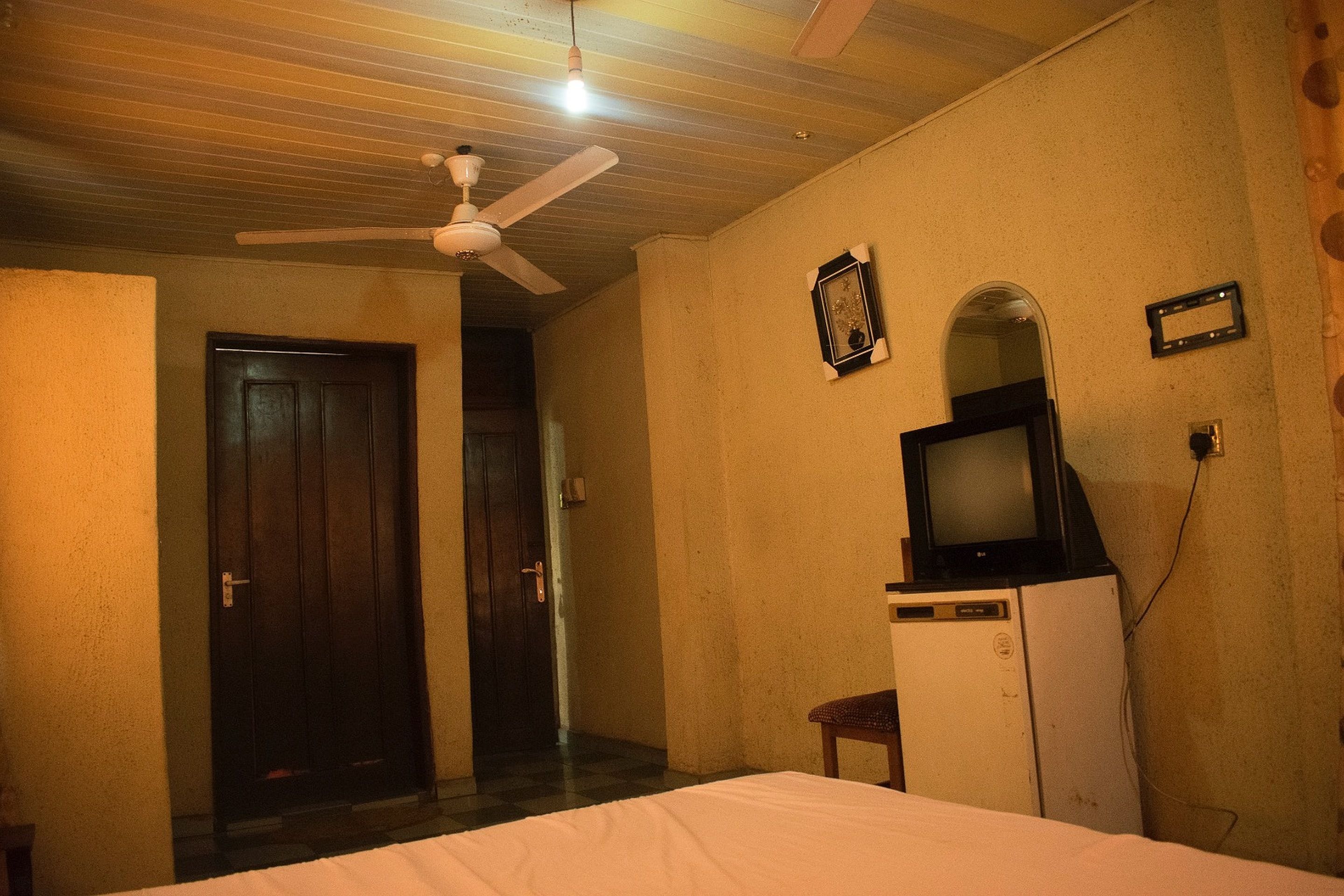 Una camera d'albergo con un letto, una ventola a soffitto, una televisione su una piccola credenza e una porta che dà su un'altra stanza.