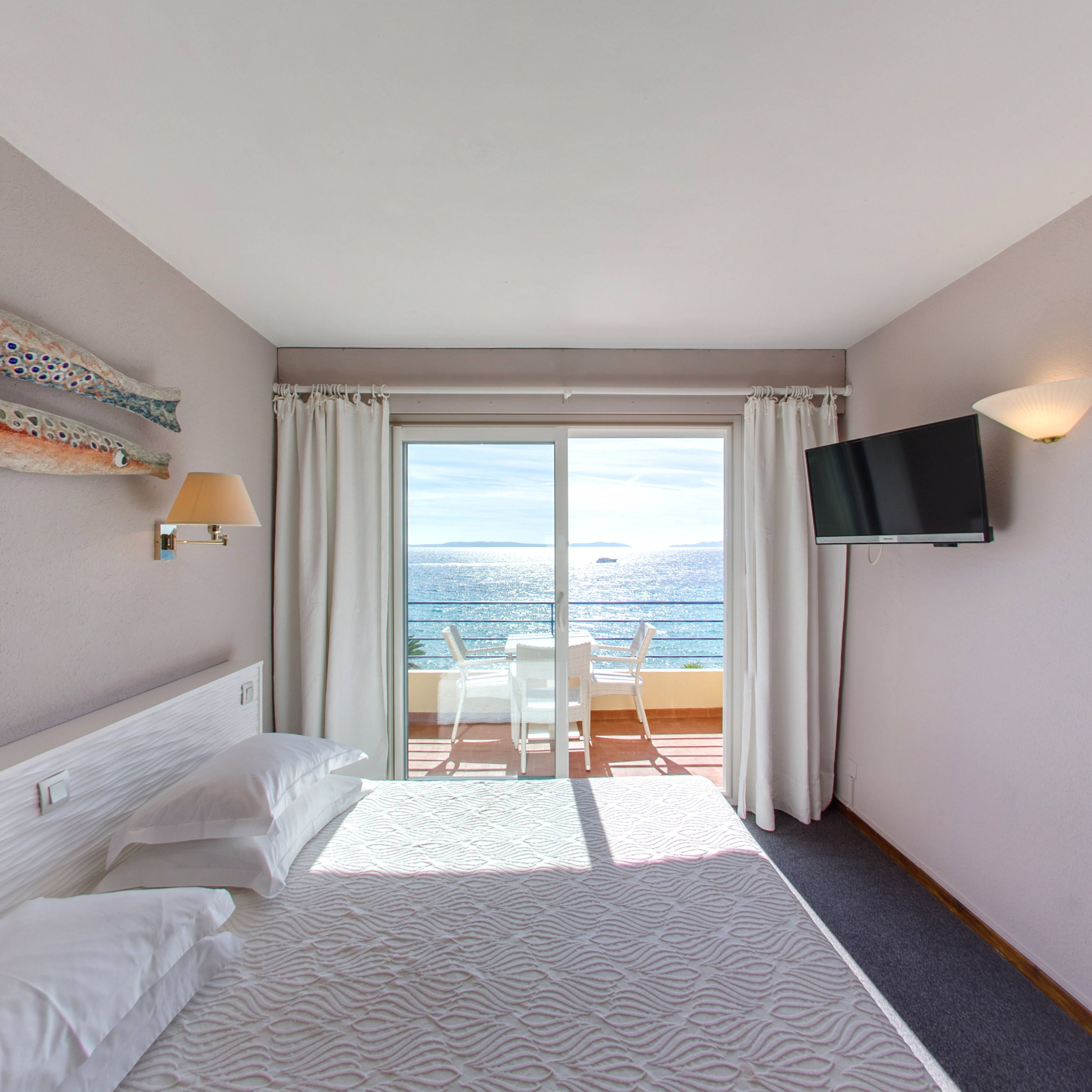 Una camera d'albergo con un letto, una TV, un balcone vista mare e una sedia.
