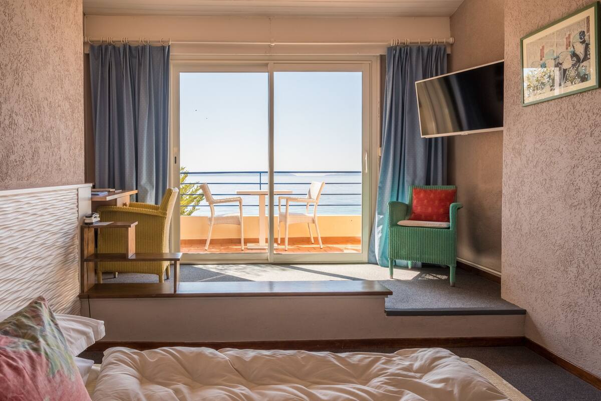 Une chambre d’hôtel équipée d’un lit, d’un bureau, d’une chaise et offrant une vue sur la mer grâce à une porte coulissante.