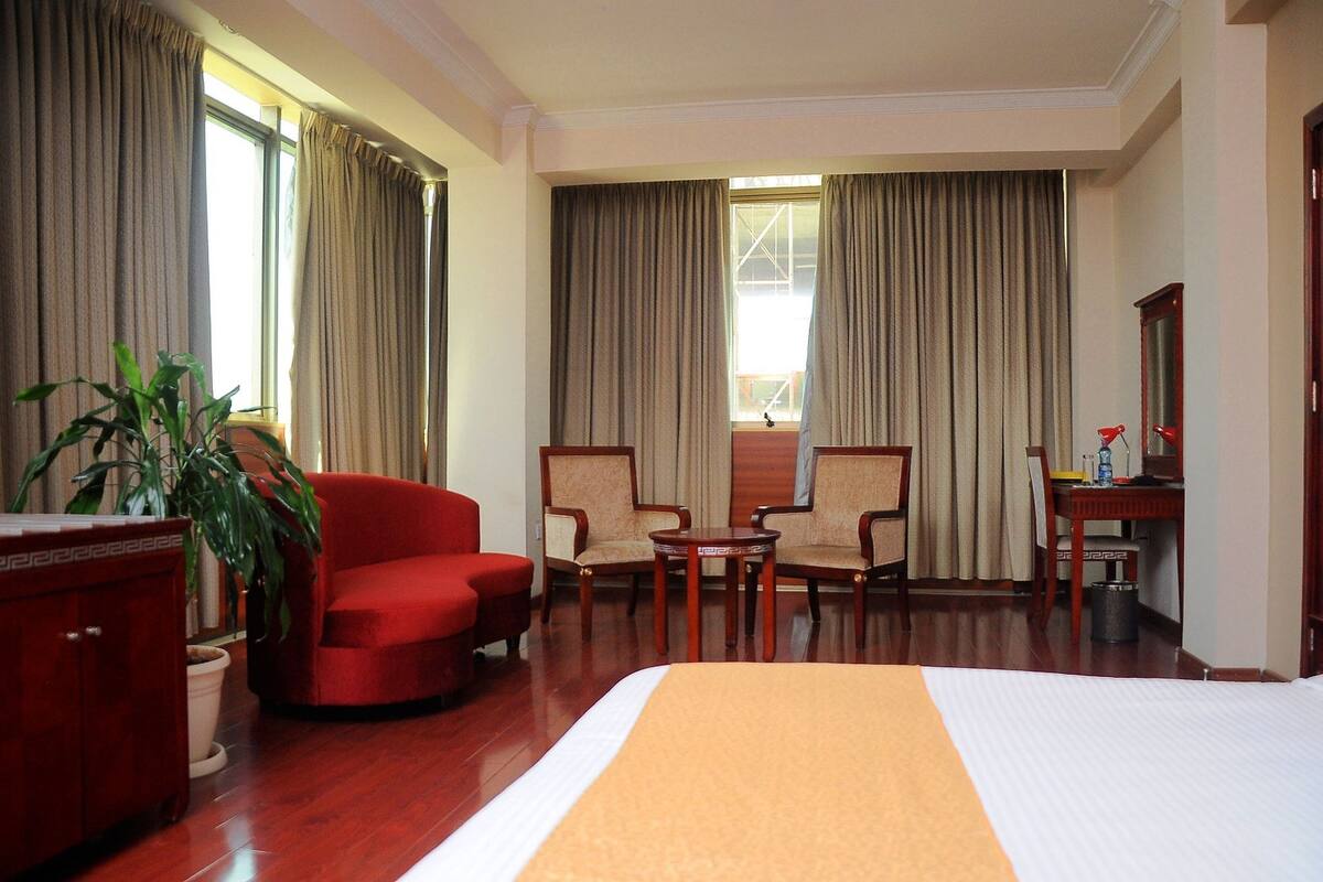 A hotel room with a large bed, two armchairs, a small table, a desk with a chair, a potted plant, and a cabinet.